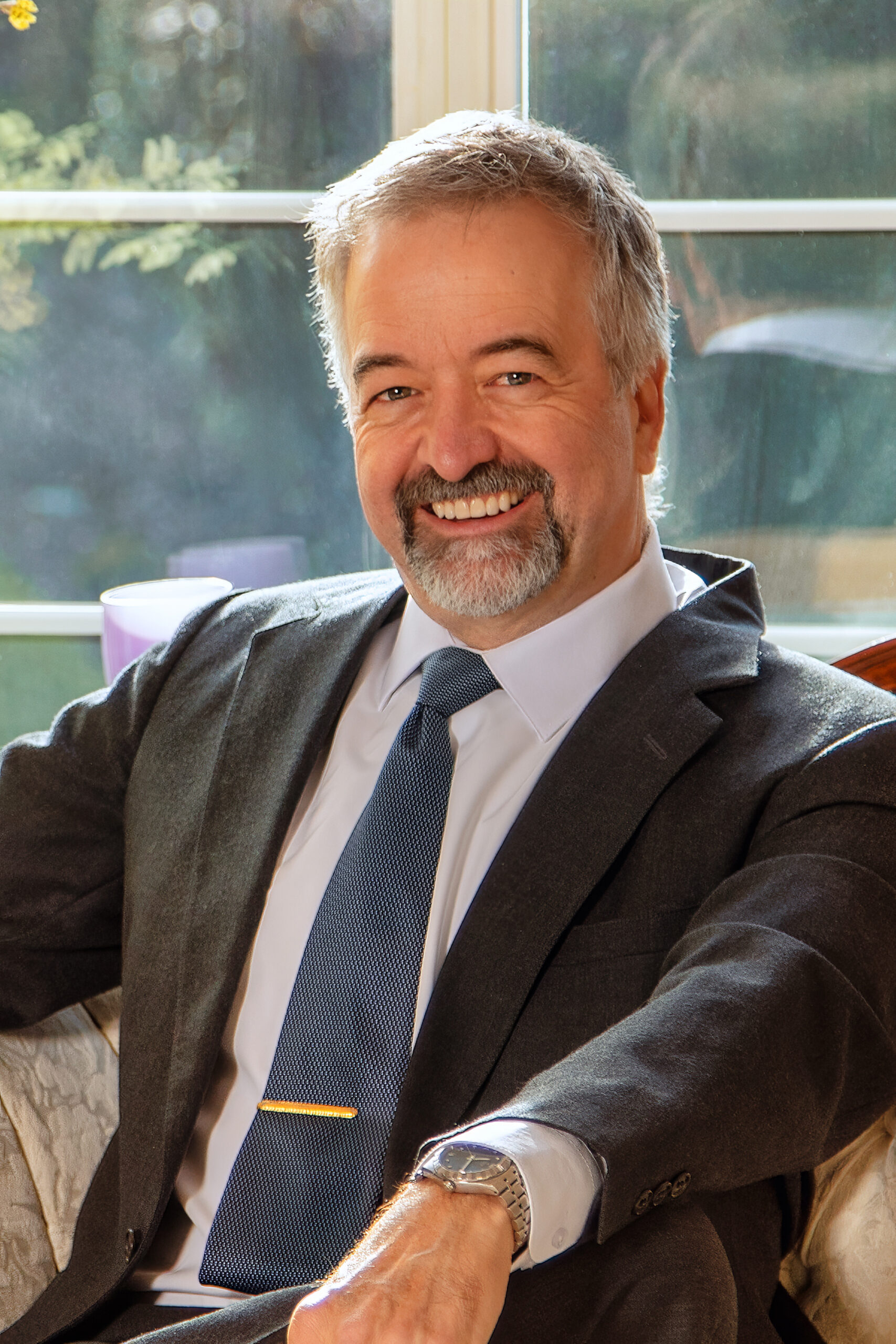 A middle-aged man with gray hair and a beard, wearing a suit and tie, sits on a couch and smiles at the camera. Sunlight streams through a window behind him. - Duvall Chamber