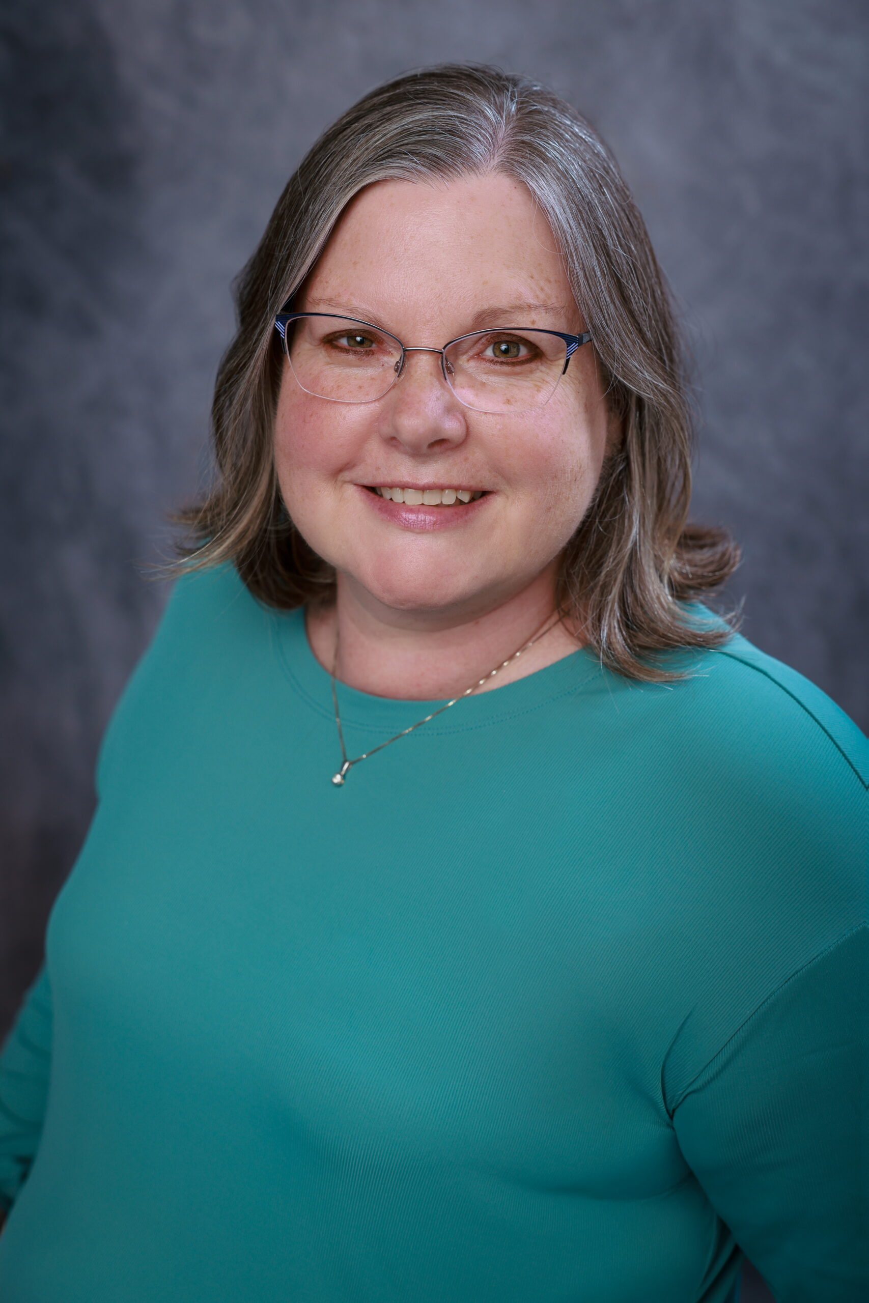 A woman with shoulder-length gray hair and glasses, wearing a teal top and a small necklace, smiles at the camera against a textured gray background. - Duvall Chamber