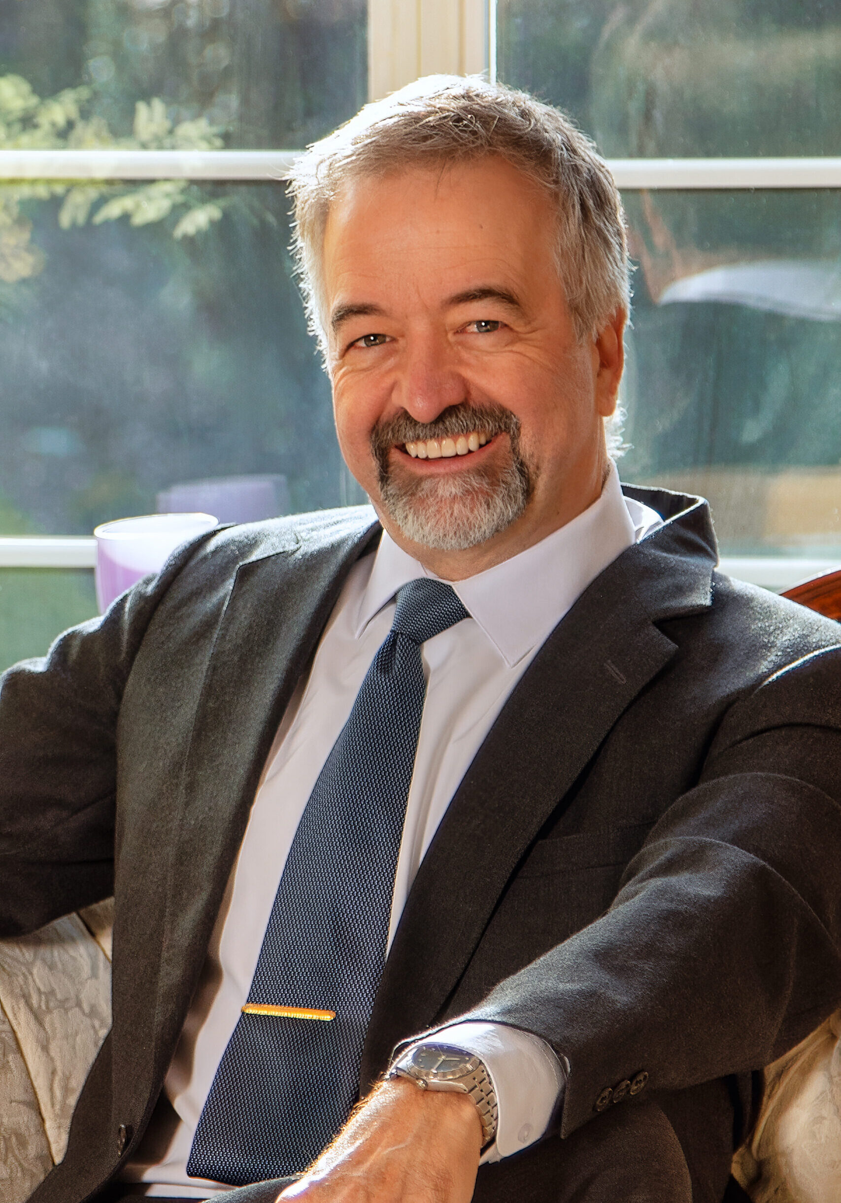 A middle-aged man with gray hair and a beard, wearing a suit and tie, sits on a couch and smiles at the camera. Sunlight streams through a window behind him. - Duvall Chamber