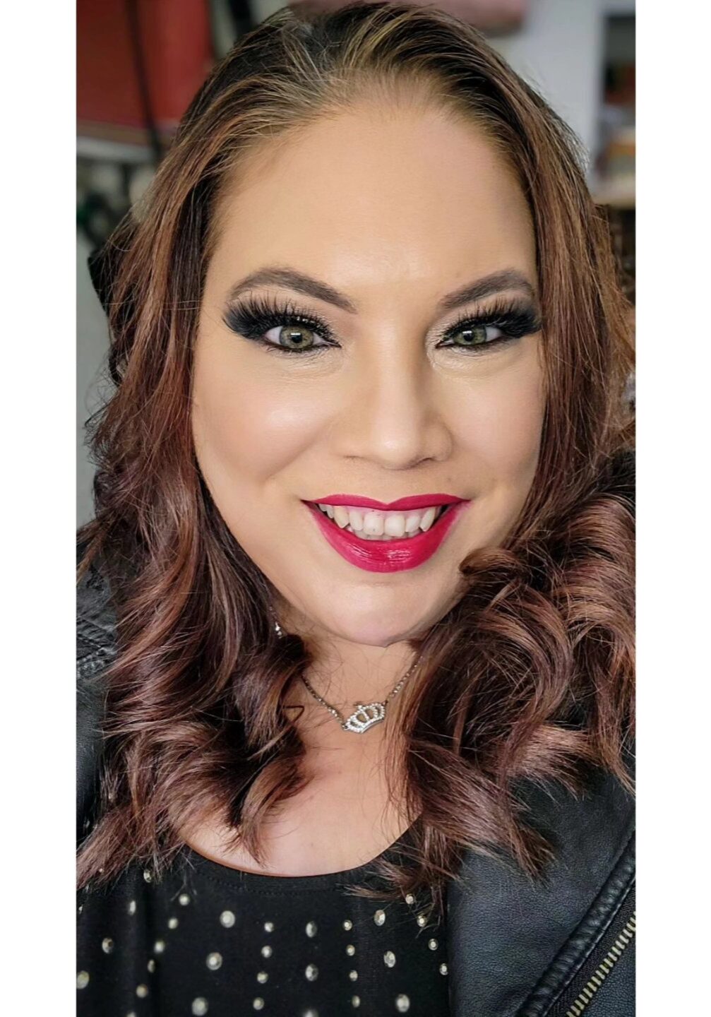 Woman with wavy brown hair, dramatic eye makeup, and red lipstick smiles at the camera, wearing a black top with studs and a necklace. - Duvall Chamber