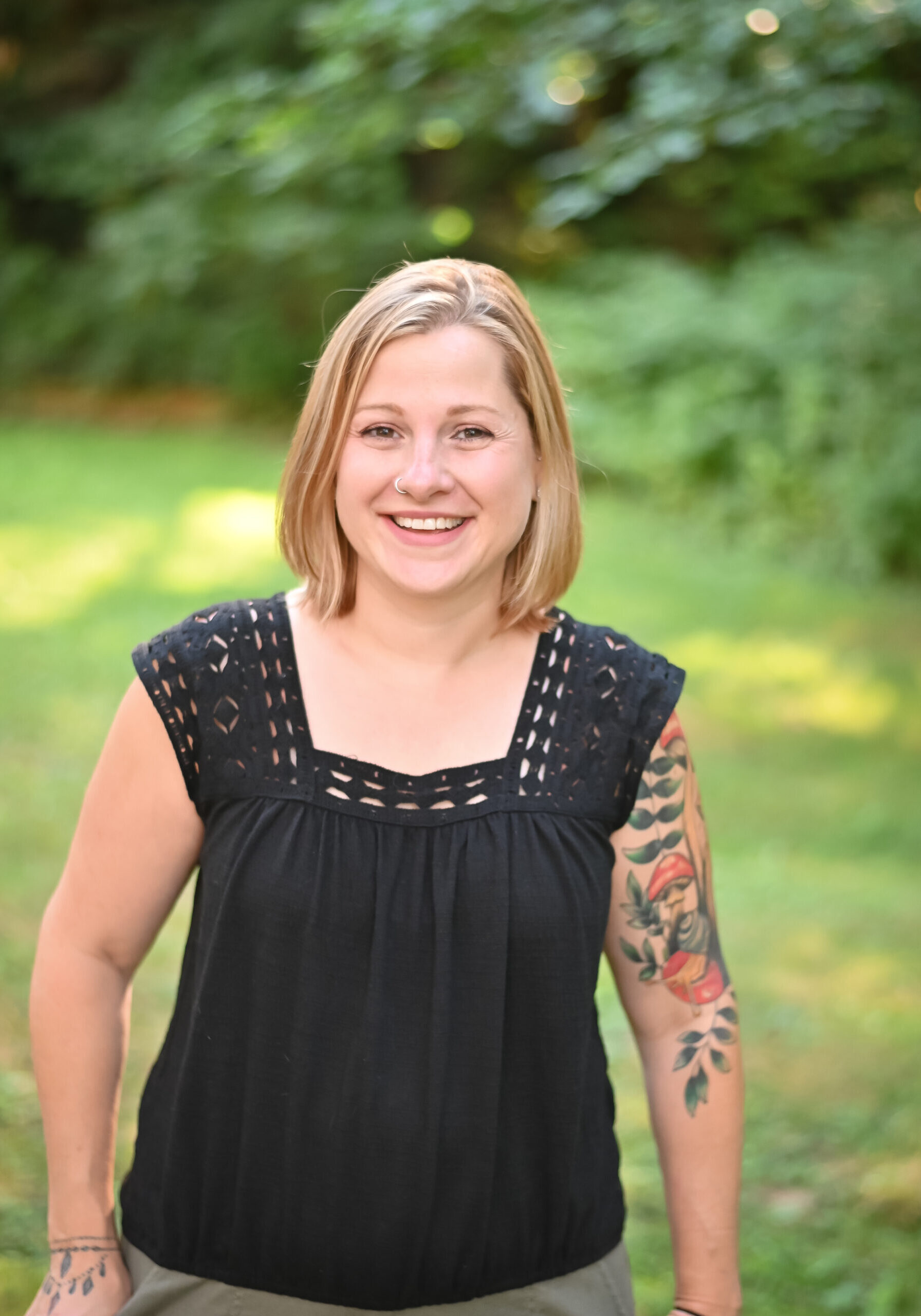 A woman with blonde hair and a floral tattoo on her left arm stands outside on grass, smiling at the camera. She is wearing a black sleeveless top. Trees and greenery are in the background. - Duvall Chamber
