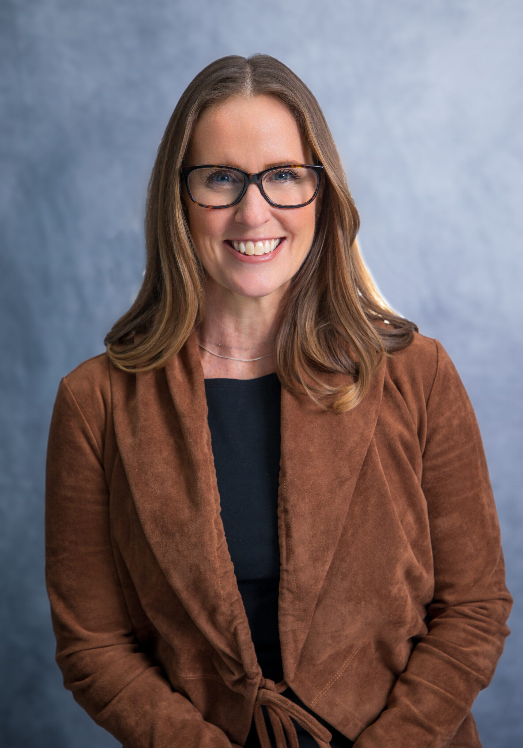 Woman with long brown hair and glasses, wearing a brown suede jacket and black top, smiling in front of a plain gray background. - Duvall Chamber