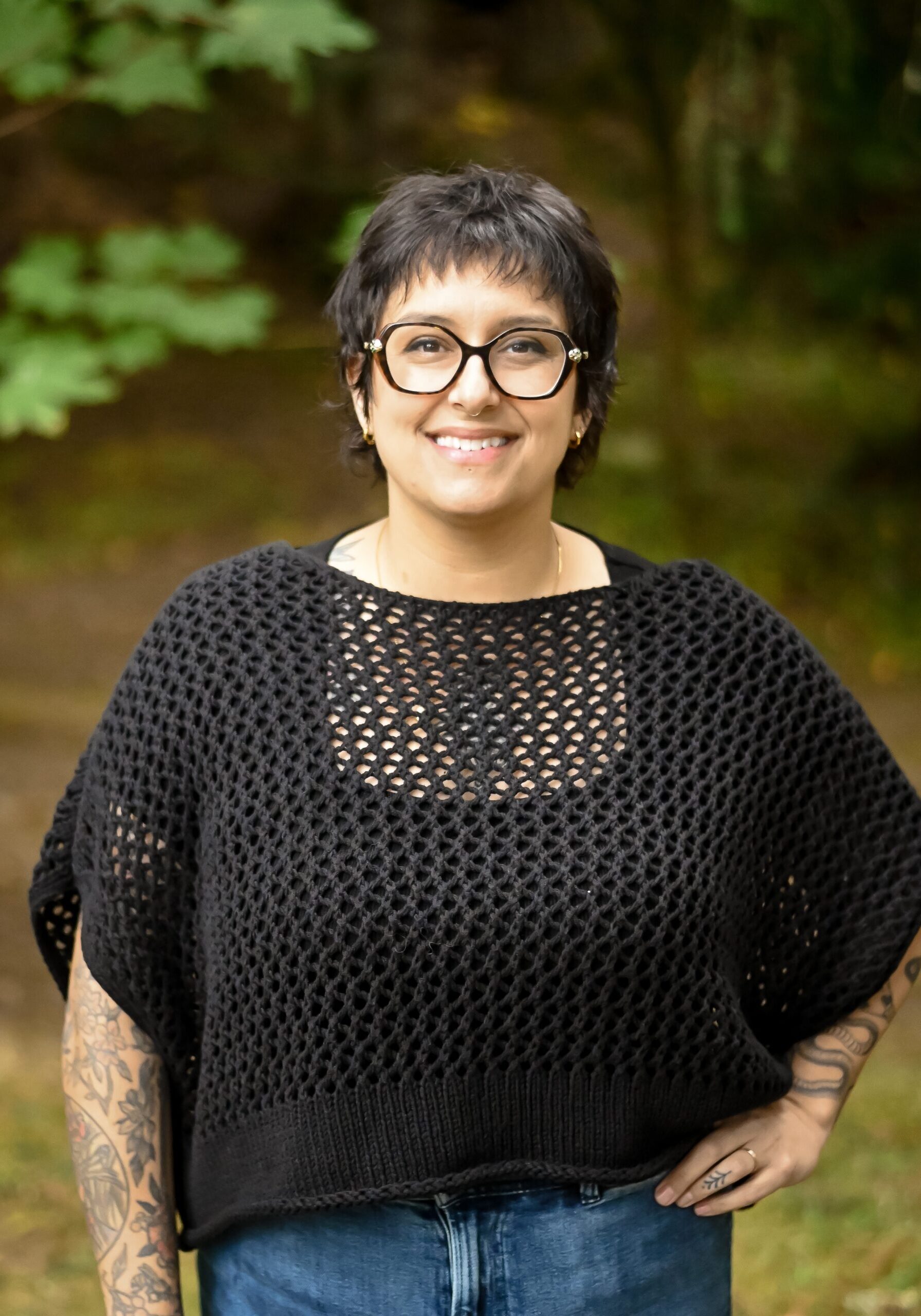 A person with short dark hair, glasses, tattoos, and a black knit top stands outdoors, smiling at the camera with one hand on their hip. - Duvall Chamber