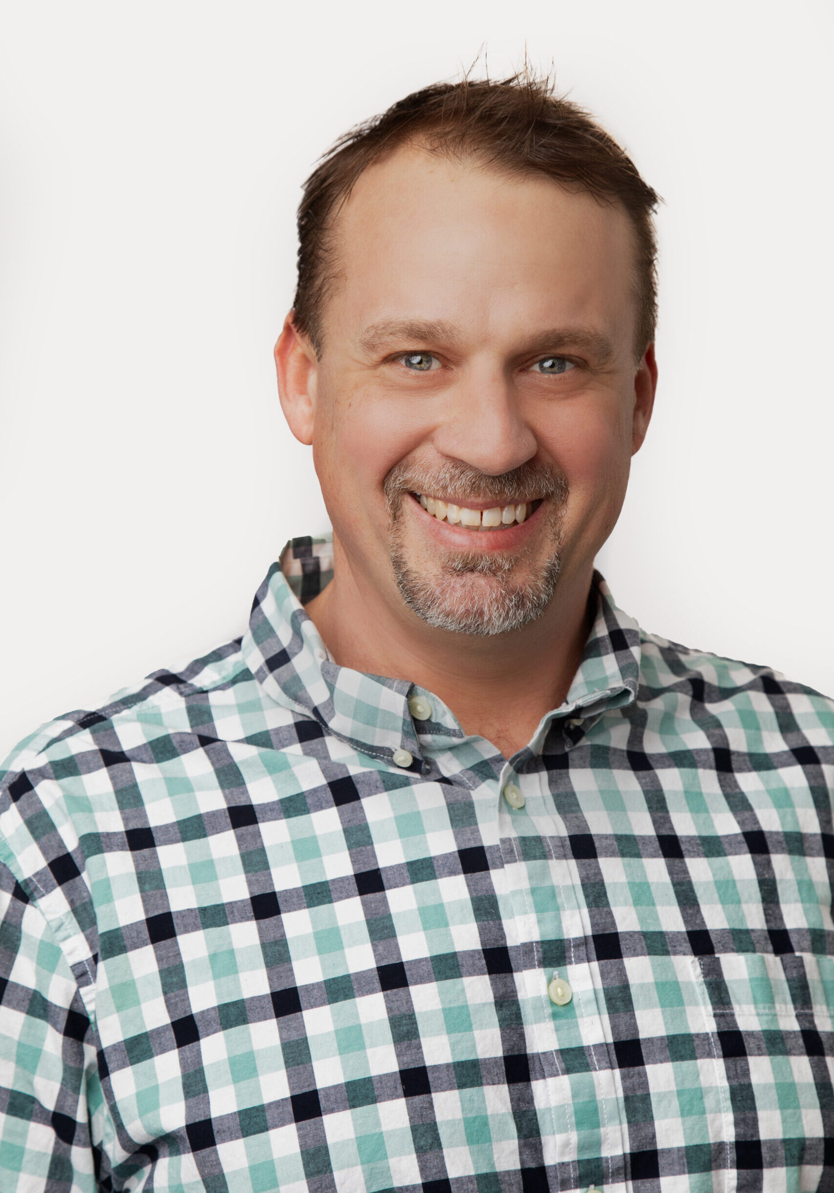 A man with short brown hair and a trimmed beard smiles at the camera, wearing a plaid shirt against a plain white background. - Duvall Chamber